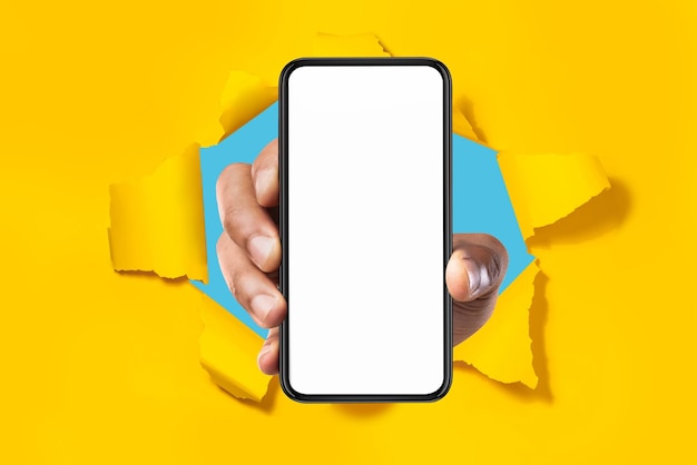 Aplicación celular Mano masculina que muestra el teléfono inteligente con pantalla blanca en blanco rompiendo la maqueta de fondo de papel amarillo roto