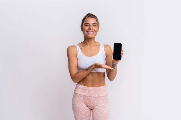 Aplicación de entrenamiento. Ajuste a una mujer bonita que muestra un teléfono inteligente con una aplicación de fitness moderna para realizar un seguimiento de la actividad deportiva. Posando sobre la pared amarilla del estudio.