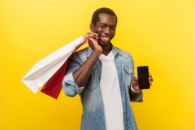 Aplicación móvil para compras en línea Retrato de un hombre positivo con una atractiva sonrisa con dientes en una camisa casual de mezclilla sosteniendo paquetes y mostrando una foto de estudio en el interior del teléfono inteligente aislada en un fondo amarillo