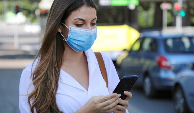 Aplicación móvil de coronavirus pandémico COVID-19 - Mujer joven con máscara quirúrgica usando la aplicación de teléfono inteligente en City Street