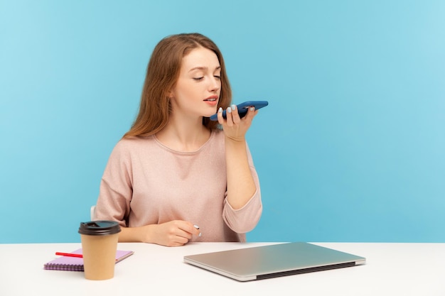 Aplicativo de alto-falante digital. empregada jovem sentada no local de trabalho e falando com o celular, usando o assistente de voz para gravar o lembrete do evento de negócios. tiro de estúdio interno isolado em fundo azul