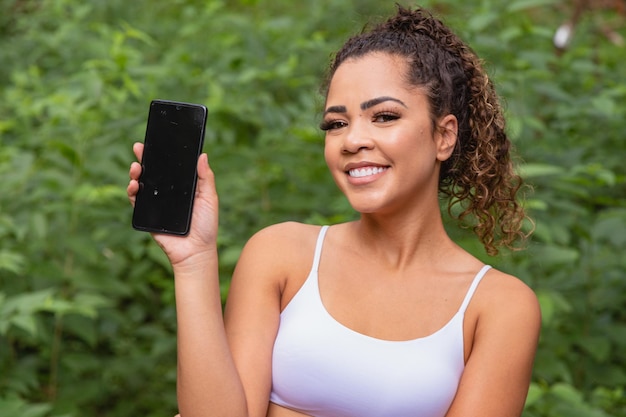 Aplicativo de esportes. Retrato de jovem apto animado segurando e mostrando o smartphone, apontando e indicando uma tela em branco para texto. Ao ar livre