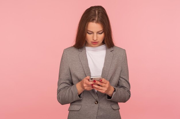 Aplicativo de negócios móveis. Retrato de chefe de senhora elegante focada séria em terno lendo mensagem, verificando notícias no smartphone, usando serviços on-line. tiro de estúdio interior isolado no fundo rosa