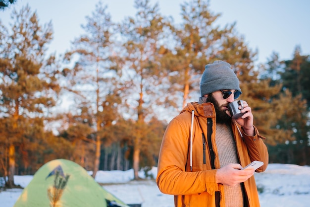 Após a subida, o turista admira o pôr do sol em pé perto da barraca com uma xícara de chá ou café e tira uma foto com o celular