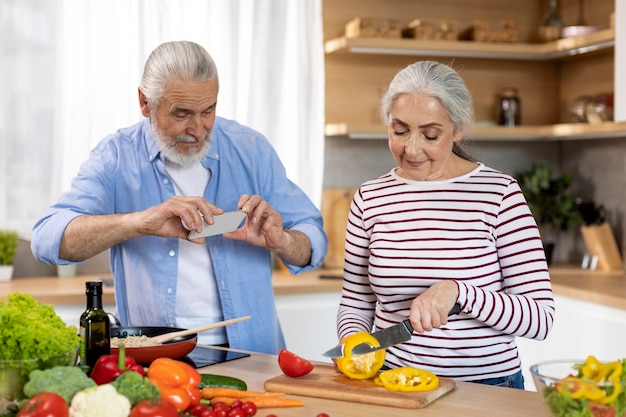 Aposentadoria hobbies homem sênior com smartphone capturando sua esposa cozinhando na cozinha