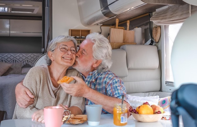 Aposentados idosos em estilo de vida alternativo dentro de uma van campista sentados tomando café da manhã juntos enquanto marido beija sua esposa