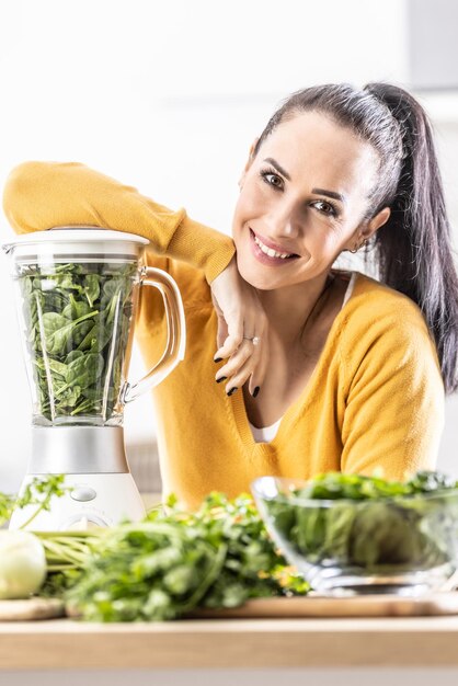 Apoyada en una licuadora, una joven sonriente prepara una bebida energética de espinacas Smoothie