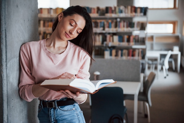 Apoyado en la pared. Chica morena en ropa casual pasando un buen rato en la biblioteca llena de libros