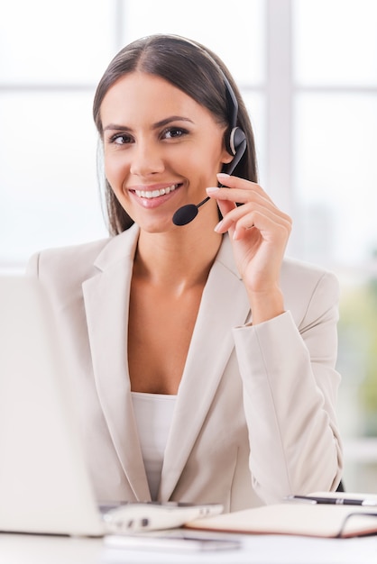 Apoyando su negocio. Hermosa joven empresaria ajustando sus auriculares y sonriendo mientras está sentada en su lugar de trabajo