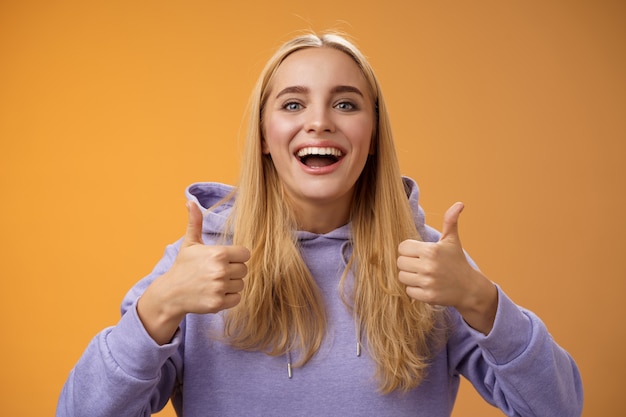 Apoyar a la linda novia rubia animando que les gusta el concepto interesante idea genial pulgar hacia arriba sonriendo ampliamente de acuerdo aprobar el atuendo de compras juntos amigos, recomendar adorar nuevo look, fondo naranja.