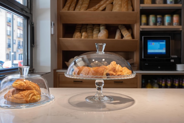 Appetitanregendes hausgemachtes Croissant oder Cruasan in einer Glastasse auf der Theke einer Konditorei, mit einem Regal im Hintergrund mit verschiedenen Brotsorten