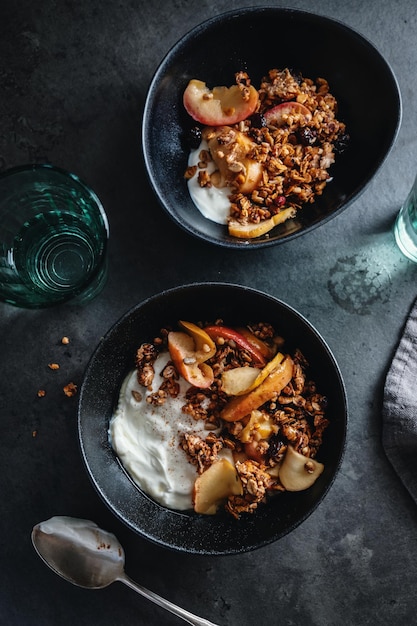 Appetitanregendes hausgemachtes herbstliches Müsli mit Äpfeln und Joghurt in Schüssel auf dunklem Hintergrund serviert.