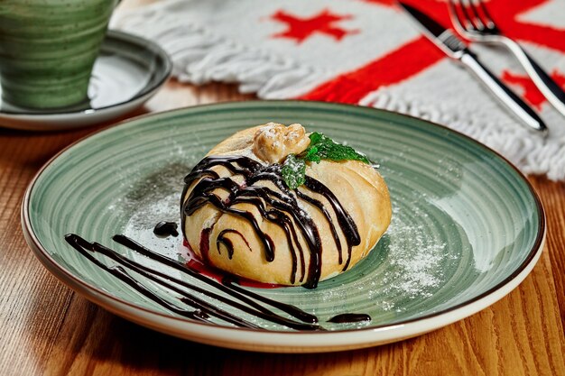 Appetitlich gebackenes Brötchen mit saftiger Beerenfüllung, bestreut mit Puderzucker, serviert mit Schokoladendressing und frischen Minzblättern im georgischen Restaurant im Hintergrund mit nationaler Five Cross Flag