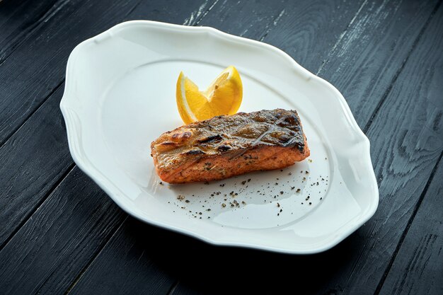 Appetitlich gegrilltes Lachssteak auf Holzkohle mit Zitrone, serviert in einem weißen Teller auf dunklem Holzhintergrund. BBQ Meeresfrüchte