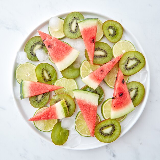 Appetitlich saftige Kiwi-, Limetten- und Wassermelonenstücke mit Eiswürfeln in einem Teller auf grauem Marmorhintergrund. Flach legen