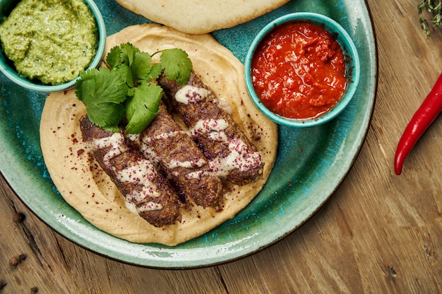 Appetitliche Brötchen Lyulya-Kebab - gekocht über einem Lammhackfleisch mit einer Beilage Hummus mit Pita und Salsa auf Keramikplatte. Vertikale Holzoberfläche, östliche Küche