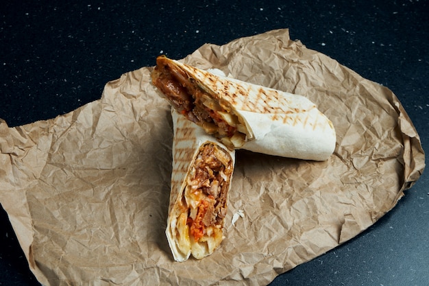 Appetitliche Dönerrolle mit Fleisch, Salat und hausgemachter Sauce in dünnem Fladenbrot auf Bastelpapier auf einem schwarzen Tisch. Östliche Küche. Geschnittener Kebab mit gegrilltem Fleisch.