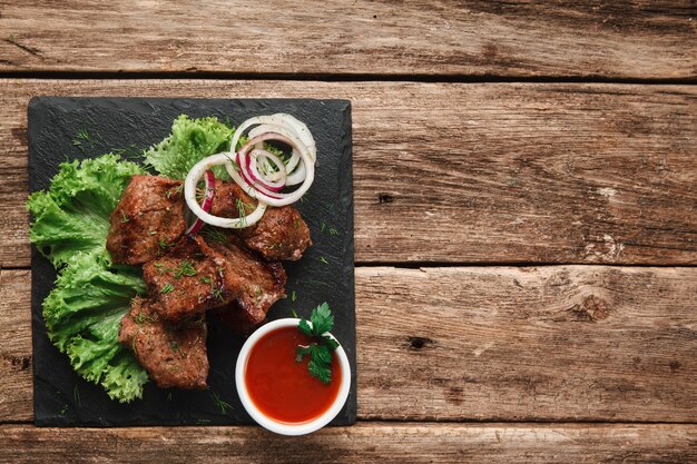 Appetitliche Grillmahlzeit. Schaschlik serviert mit Salat und Zwiebelringen auf schwarzem Slade mit Tomatensauce, auf rustikalem Holztisch mit Freiraum.