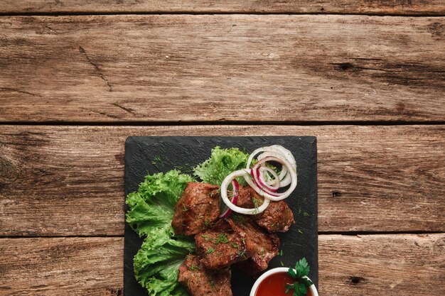 Appetitliche Grillmahlzeit. Schaschlik serviert mit Salat und Zwiebelringen auf schwarzem Slade mit Tomatensauce, auf rustikalem Holztisch mit Freiraum.