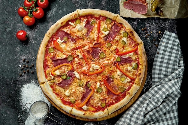 Appetitliche italienische Pizza mit Käse, Paprika, Schinken, eingelegten Gurken und roter Sauce in einer Komposition mit Zutaten auf einem schwarzen Tisch