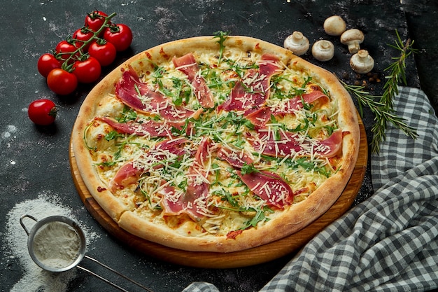 Appetitliche italienische Pizza mit Käse, Schinken, Rucola und Parmesan in einer Komposition mit Zutaten auf einem schwarzen Tisch