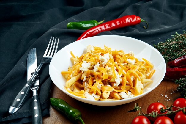 Appetitliche Pommes Frites mit Cheddar-Käse in einer weißen Schüssel auf einem Holztisch in einer Zusammensetzung mit Zutaten. Pommes Frites mit Belag