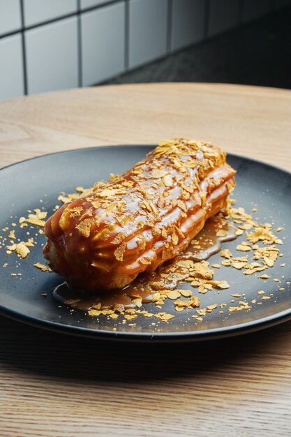 Appetitlicher und großer Eclair mit Glasur aus gesalzenem Karamell und Tannencreme auf einem schwarzen Teller. Süßigkeiten und Dessert für Kaffee und Tee.