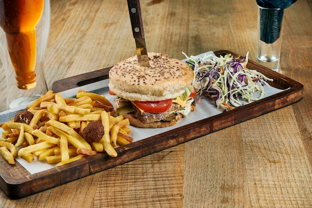 Appetitlicher und saftiger Burger mit Rindfleischpastetchen, geschmolzenem Cheddar-Käse, Tomaten und Zwiebeln auf einem weißen Teller mit Pommes Frites. Nahansicht. Holzoberfläche