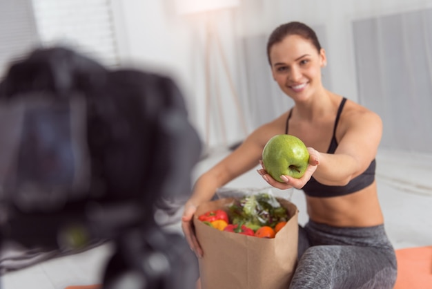 Apple para la salud. Mujer joven atlética bastante alerta sonriendo y con un paquete con verduras y mostrando una manzana mientras hace un video para su blog