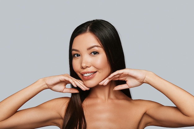 Apreciando sua beleza. Mulher jovem e bonita asiática olhando para a câmera e sorrindo em pé contra um fundo cinza