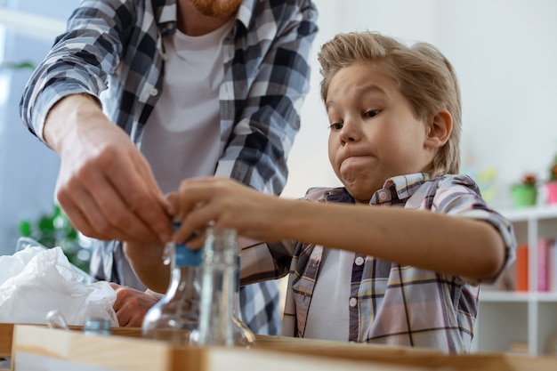 Aprendendo a classificação cuidadosa. Garoto loiro concentrado observando os movimentos de sua professora enquanto lida com diferentes lixos