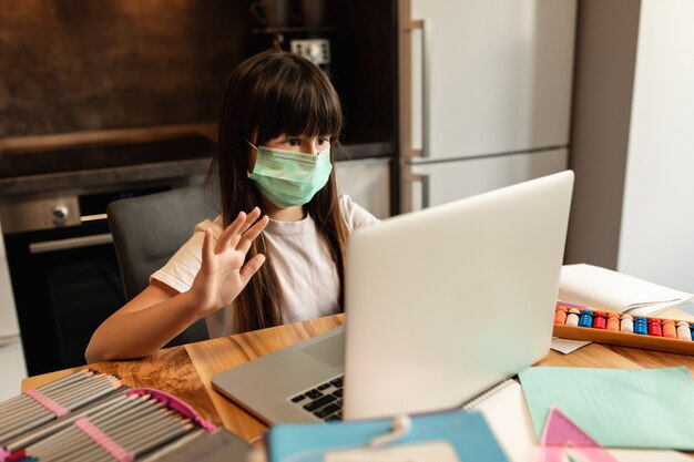 Aprendizado on-line durante a quarentena. Menina com uma máscara protetora no rosto fala com outros participantes na aula de vídeo on-line. Educação on-line em casa.