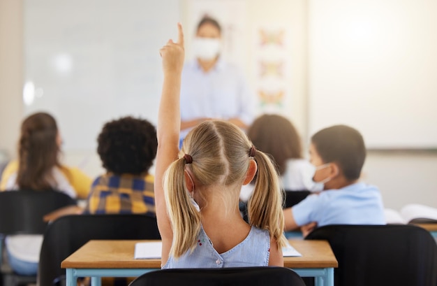 Aprendizagem inteligente e garotinha na classe segurando uma mão para responder a uma pergunta na escola Vista traseira de um jovem estudante sentado em uma mesa de sala de aula olhando para resolver as perguntas que um professor está fazendo