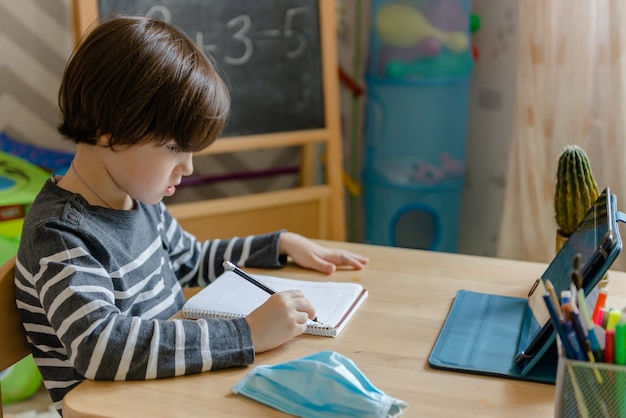 Aprendizaje a distancia para niños durante la epidemia de coronavirus. Un niño se sienta en una mesa y realiza tareas de profesor en Internet.