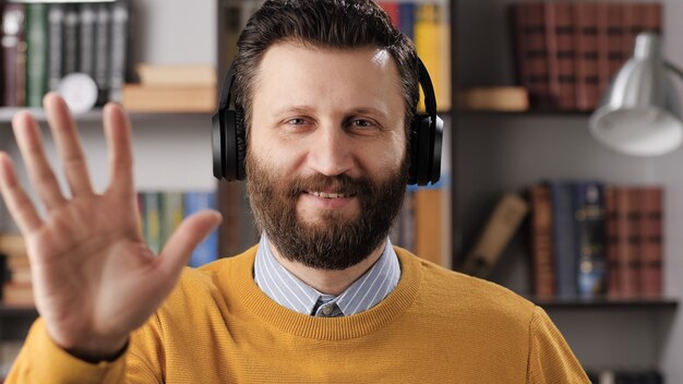 Aprendizaje en línea, educación remota, videollamada, e-learning, concepto de presentación remota. Hombre barbudo positivo mirando a cámara y muestra HOLA con su mano. Tiro medio