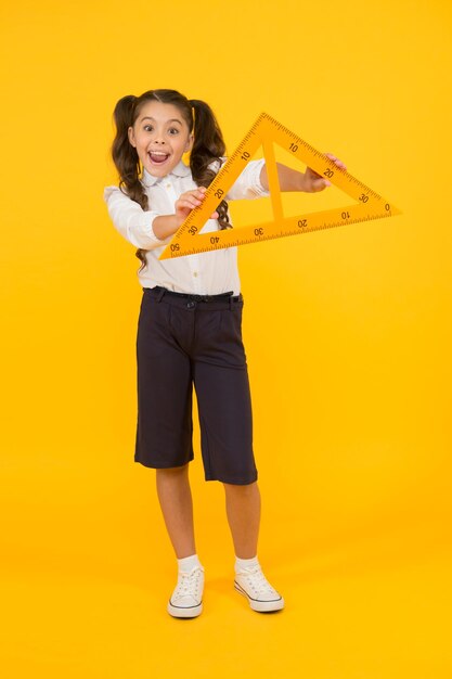 Aprendo geometría fácilmente Adorable niño pequeño aprende geometría en la escuela Linda niña pequeña sosteniendo un triángulo para aprender matemáticas sobre fondo amarillo Estar ansioso por aprender