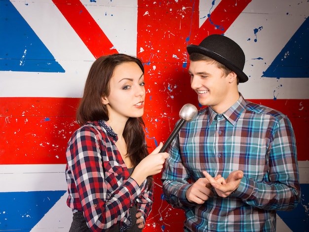 Apresentadora de um programa de bate-papo britânica entrevistando um jovem bonito com um chapéu-coco em frente a uma bandeira da Union Jack pintada na parede