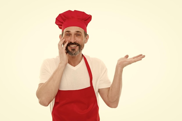 Apresentando o produto Homem maduro barbudo com chapéu de chef e avental apontando a mão de lado Cozinheiro sênior com barba e bigode usando avental de babador Cozinheiro chefe maduro no espaço de cópia do avental de cozinha vermelho