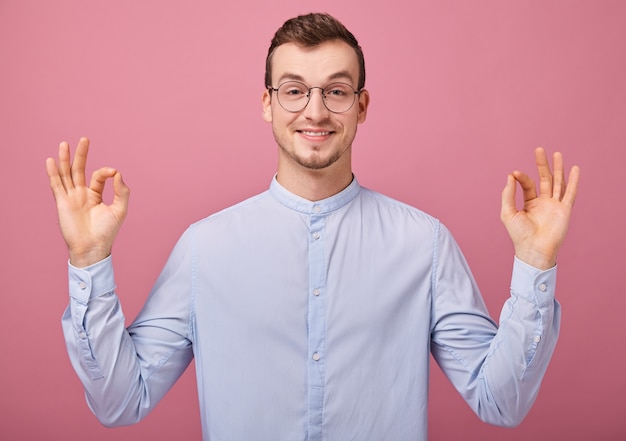 Foto apresentável jovem de camisa azul e óculos aponta o dedo para a direita, sorrindo