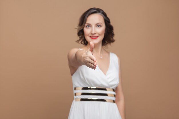 Apretón de manos. mujer feliz sonrisa con dientes y mostrando el cartel de bienvenida. Mujer de expresión emocional en vestido blanco, labios rojos y peinado rizado oscuro. Foto de estudio, interior, aislado sobre fondo marrón beige