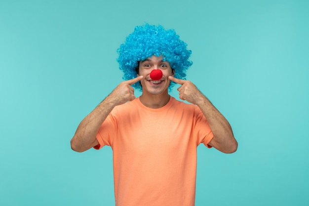 Aprilscherzkerl clown glücklich zeigt mit dem finger auf wangen lächelnd blaue haare aufgeregt lustige rote nase