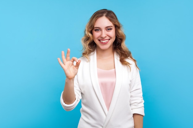 Aprovação de sinal ok Retrato de mulher adorável feliz com cabelos ondulados em jaqueta branca casual, fazendo um gesto bem e sorrindo com alegre expressão satisfeita satisfeita estúdio interior tiro fundo azul