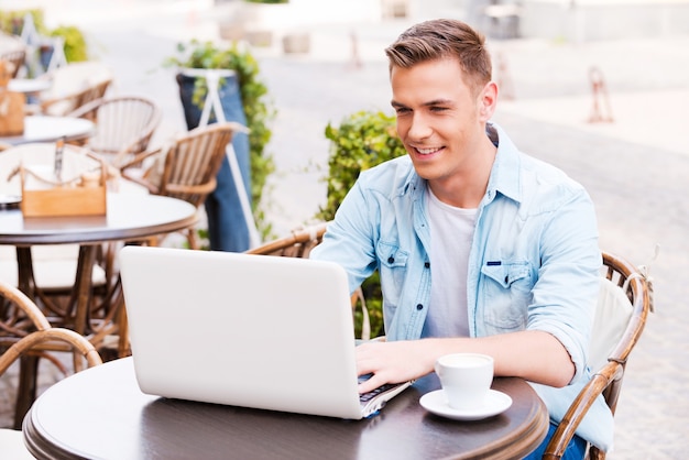 Aprovechar las ventajas de la conexión Wi-Fi gratuita. Apuesto joven trabajando en una computadora portátil y sonriendo mientras está sentado en la cafetería en la acera