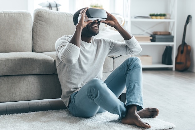 Aproveitando a nova realidade! Africano jovem e bonito ajustando seu fone de ouvido VR enquanto está sentado no tapete em casa