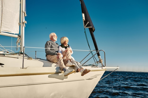 Aproveitando as férias, um feliz casal de idosos sentado ao lado de um barco a vela ou no convés de um iate