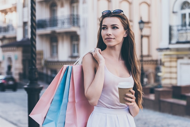 Aproveitando o café após um dia de compras. Mulher jovem e bonita carregando sacolas de compras e segurando a xícara de café enquanto caminha pela rua