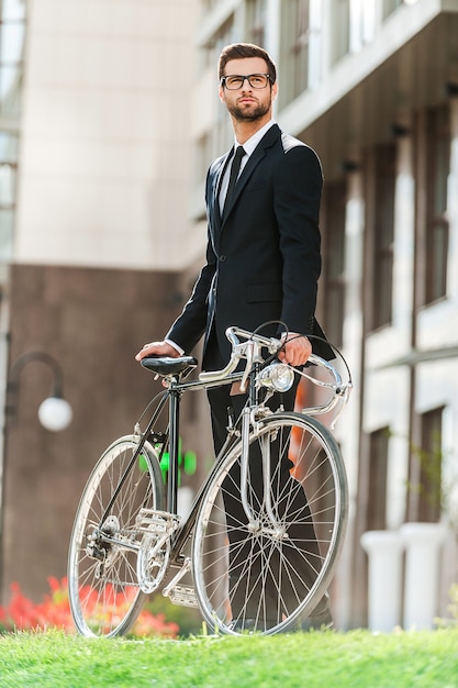 Aproveitando o estilo de vida urbano. Vista de baixo ângulo do jovem empresário confiante olhando para longe e segurando a bicicleta de mãos dadas ao ar livre