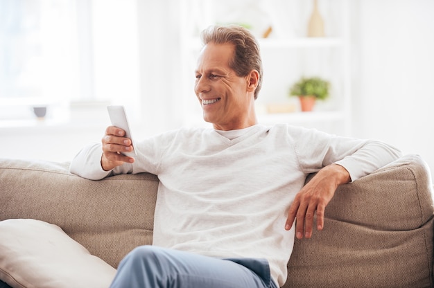 Aproveitando o tempo em casa. Homem maduro e alegre segurando um telefone celular e olhando para ele com um sorriso enquanto está sentado no sofá em casa