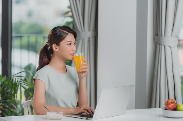 Aproveitando o tempo em casa Linda jovem sorridente trabalhando no laptop e bebendo suco de laranja