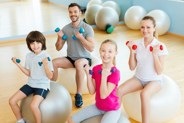 Aproveitando o tempo em uma espécie de clube. vista superior de uma família feliz e desportiva, exercitando-se com halteres no clube desportivo, sentados juntos nas bolas de fitness